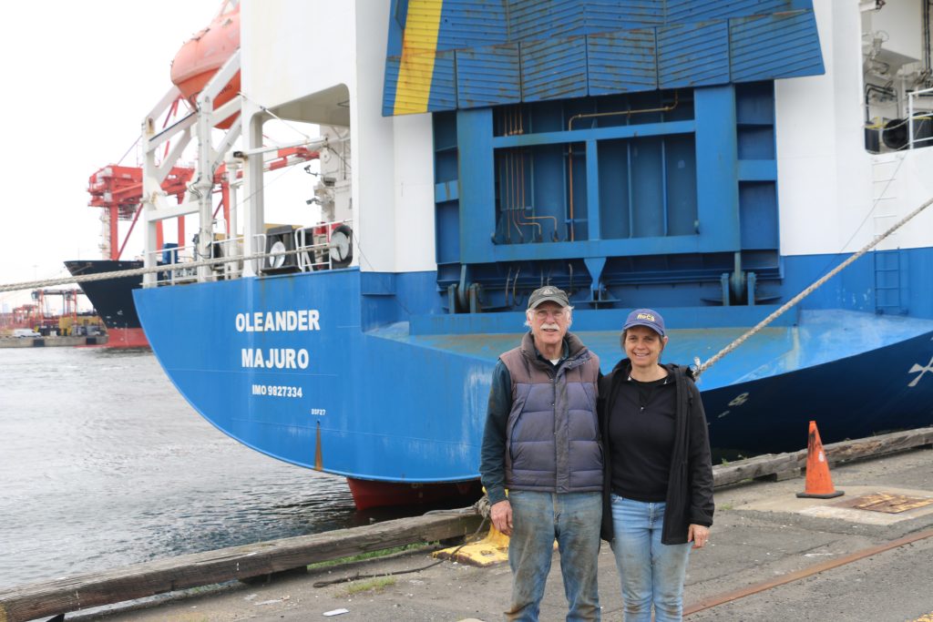 Two scientists stand in front of the stern of a ship with the name Oleander in white visible on the ship.